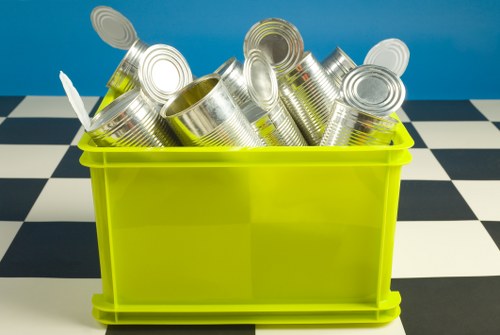 A bright yellow plastic container filled with numerous empty, silver, and metallic tin cans, some with visible ridges and open tops, placed on a black and white checkered surface against a solid blue background. The cans are stacked and leaning slightly in various directions within the container, which is positioned in a tidy, indoor environment. The image illustrates waste or rubbish typical for collection and clearance services, aligning with garden clearance and rubbish removal themes relevant to Battersea. Garden Clearance Battersea specializes in clearing household or garden waste, including metal containers and cans, from local properties. The overall scene emphasizes the handling of metallic waste, prepared for recycling or disposal, within a clean, controlled setting suitable for rubbish removal operations.