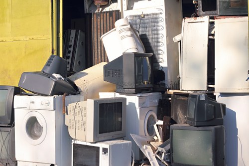 A collection of discarded household appliances and electronic waste stacked together outdoors, including washing machines, microwave ovens, and refrigerators, with some items showing signs of dirt and weathering. The appliances are arranged haphazardly on a paved surface, with a corrugated metal wall and a yellow-painted surface visible in the background, suggesting a waste disposal or recycling site. The appliances display various materials such as metal, plastic, and glass, with finishes ranging from smooth plastic to textured metal surfaces, primarily in shades of white, black, and grey. Some of the appliances are upright, while others are tilted or leaning, and a few have doors or panels partly open, revealing internal components. The overall scene looks cluttered, indicative of a rubbish removal process conducted by a professional service like Garden Clearance Battersea, servicing areas around Battersea and the broader London borough. The lighting appears natural, casting soft shadows, emphasizing the array of waste ready for collection and recycling.