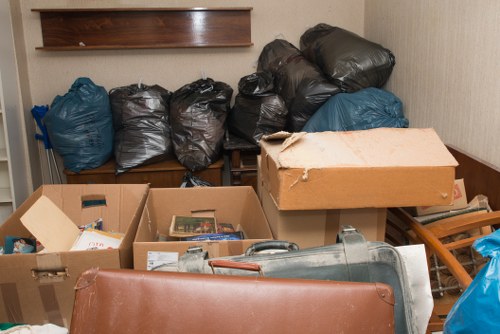 A collection of black and blue rubbish bags filled with waste, stacked on a wooden bench against a plain beige wall, with some cardboard boxes and a rolled-up carpet or mat in the foreground. The cardboard boxes contain miscellaneous items, possibly magazines or papers, and are partially open. Part of a wooden chair or small table is visible on the right side, and a grey vacuum cleaner or similar device is partially seen towards the bottom right. The scene appears to be an indoor or outdoor storage area used for rubbish collection, typical of garden clearance or waste removal services in Battersea. The lighting is neutral, highlighting the texture of the bags and cardboard, and the overall environment suggests a cleanup operation or preparation for rubbish disposal, aligning with services offered by Garden Clearance Battersea in the local postcode area.